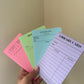 A hand holds four pastel-colored notepad sheets fanned out against a beige wall corner. The notepads include pink, green, light blue, and lavender, each with printed stationery labels such as Guest Cheque, Receipt, Notes, and Library Card.