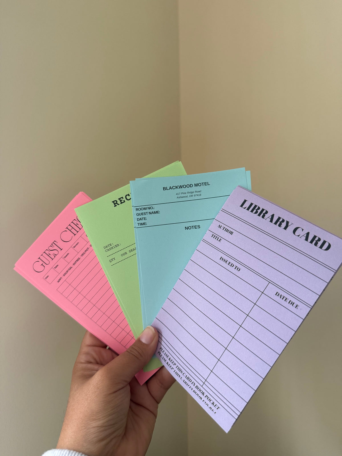 A hand holds four pastel-colored notepad sheets fanned out against a beige wall corner. The notepads include pink, green, light blue, and lavender, each with printed stationery labels such as Guest Cheque, Receipt, Notes, and Library Card.